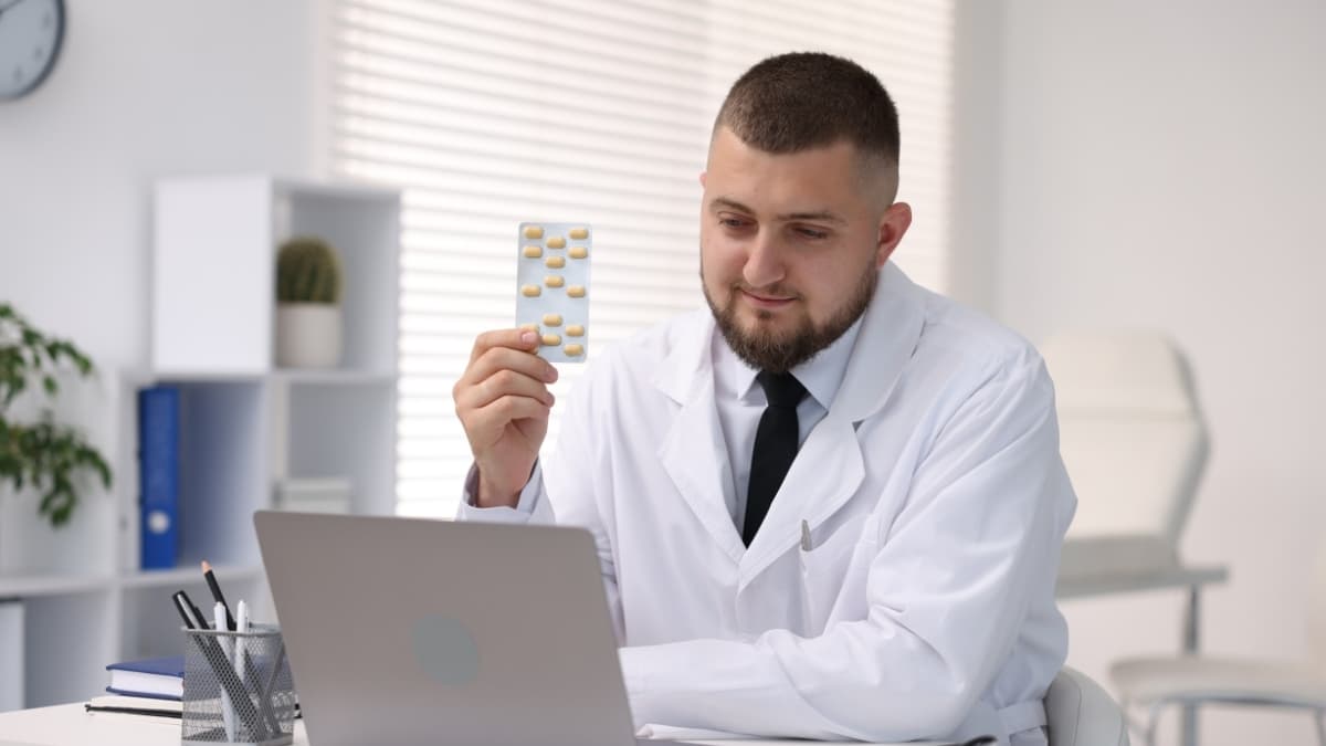 Doctor holding blister pack during online prescription consultation on laptop in clinic office