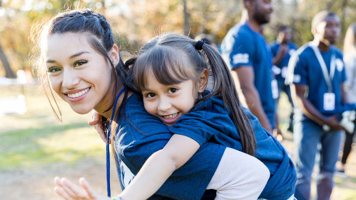 Volunteer carrying smiling young girl on her back during community charity event outdoors