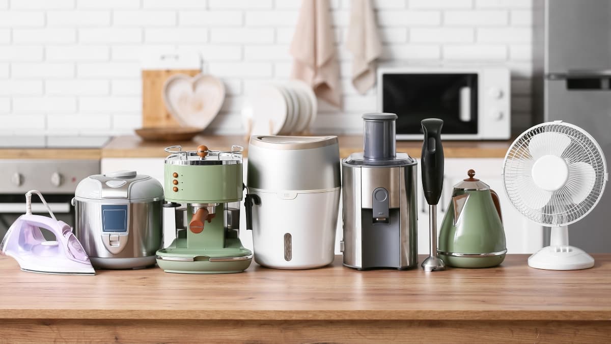 Assorted small kitchen appliances arranged on a wooden countertop including an iron, rice cooker, espresso machine, air fryer, juicer, hand blender, electric kettle, and table fan in a modern kitchen setting.