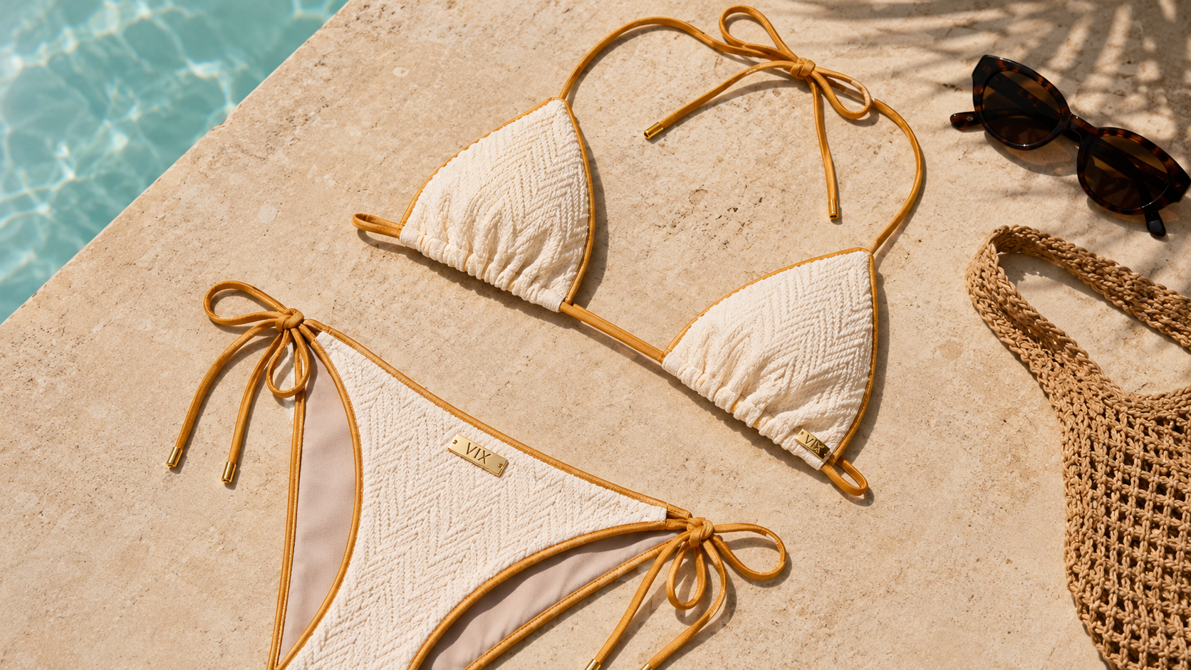 ViX Bia bikini in ivory textured fabric with gold trim, displayed on a sunlit poolside stone surface alongside sunglasses and a woven beach bag, creating a luxury resort-inspired swimwear scene.