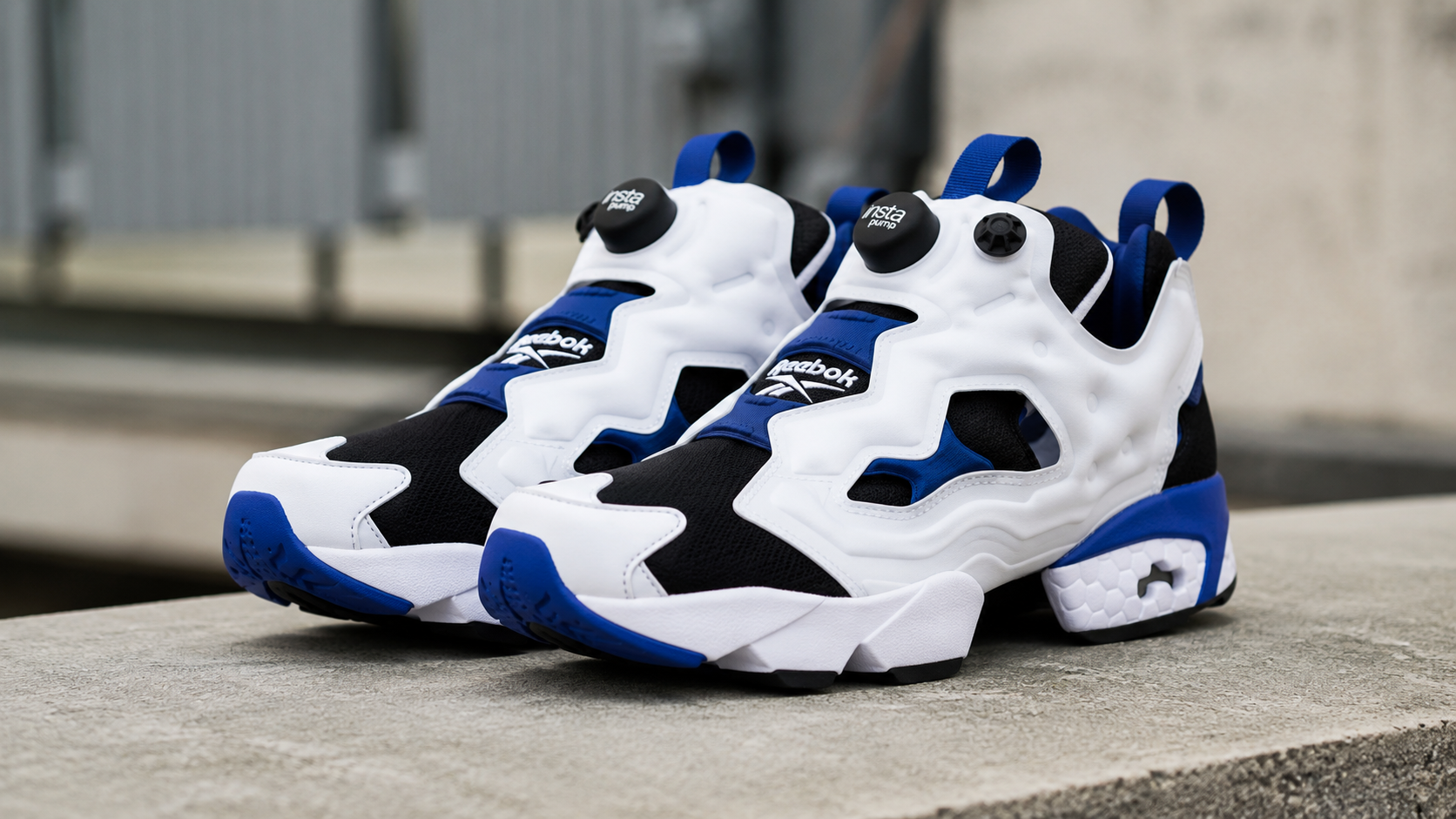 Reebok Instapump Fury 94 sneakers in white, black, and blue displayed on a concrete surface with an urban background, showcasing the iconic Pump technology and bold retro running design.