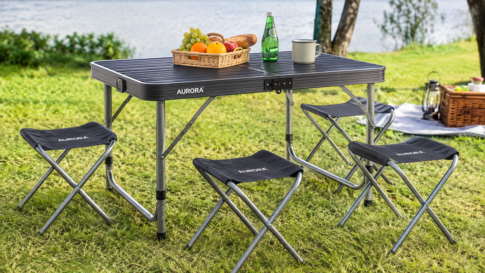 AURORA foldable picnic table and four portable stools set up on grass beside a lake with picnic essentials, ideal for camping, outdoor dining, picnics, and family gatherings.