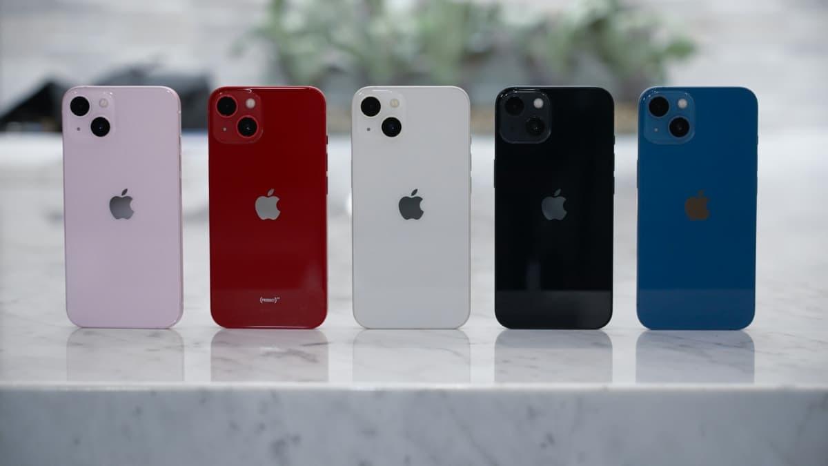 Five Apple iPhone 13 models in pink, red, Starlight, black, and blue stand upright on a marble surface.