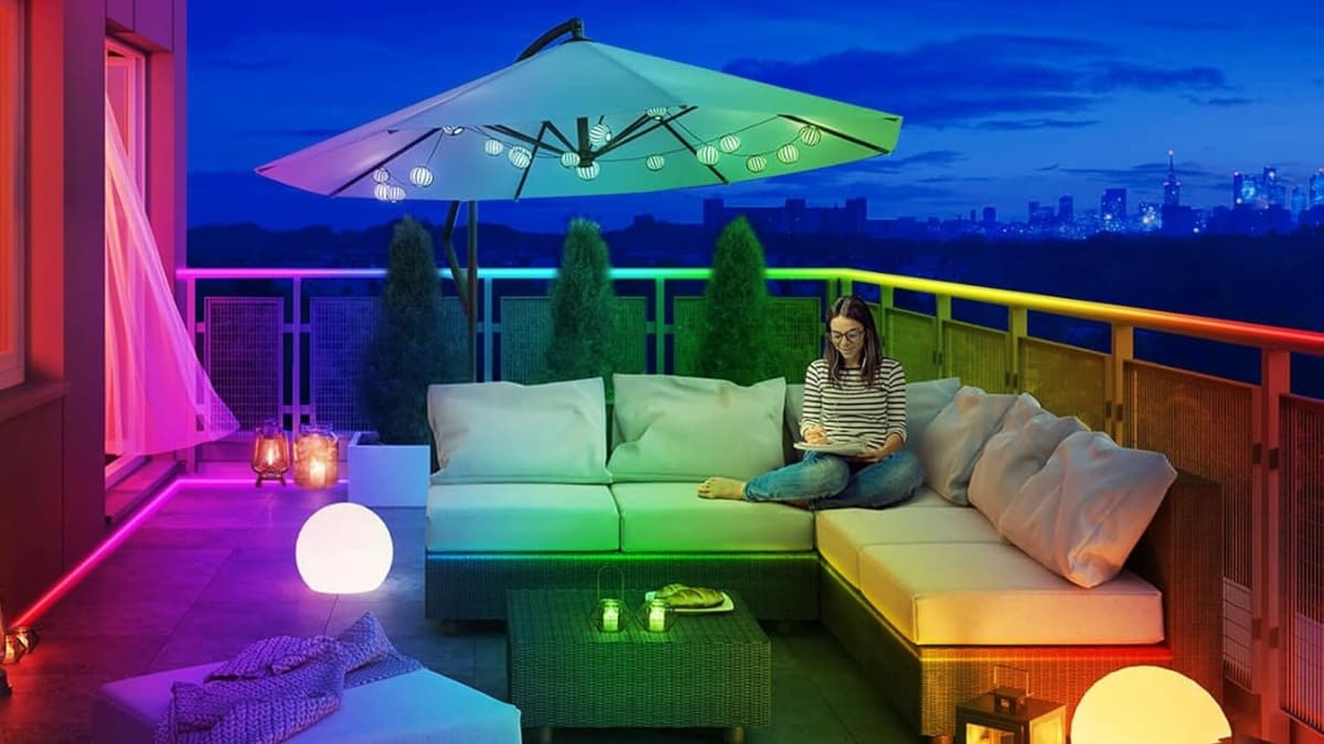 Colorful outdoor LED strip lights glow around a balcony lounge with seating, lanterns, and a large umbrella.