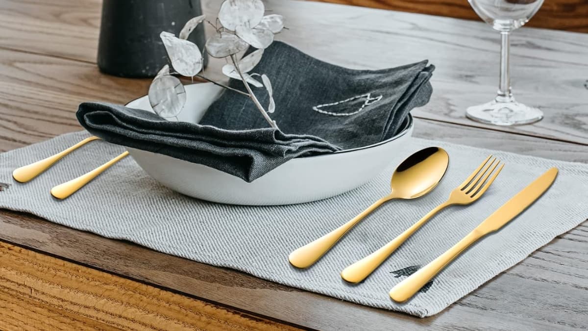 Gold silverware set placed on a gray placemat beside a white bowl with a dark napkin and decorative stem.