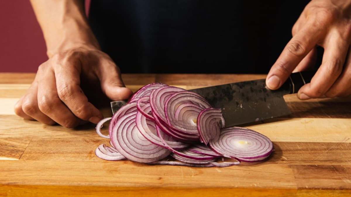 Hands slicing a red onion into thin rings on a wooden cutting board with a kitchen knife.