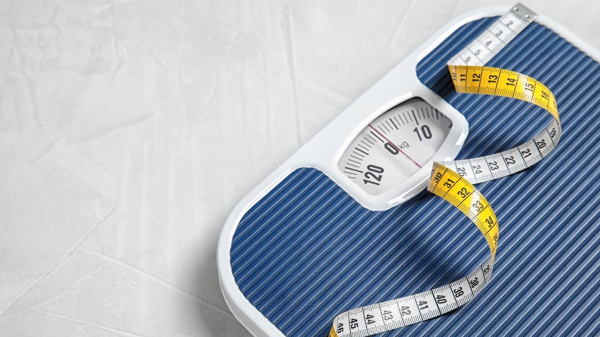 A blue analog bathroom scale with a yellow and white measuring tape placed on top, symbolizing weight tracking and body composition monitoring.