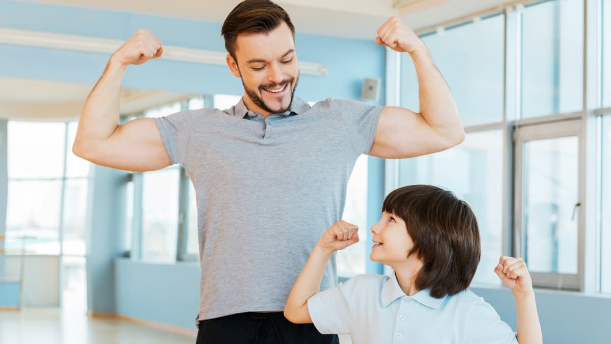 Adult showing a flexed arm pose while a child mimics the same pose during a fun workout moment at a gym.