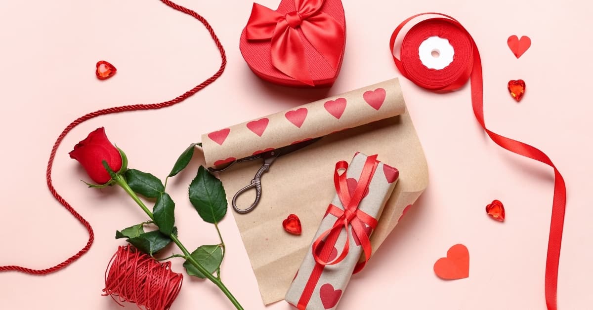 Valentine’s Day gift wrapping with red ribbon, rose, heart-shaped box, and wrapped presents on a pink background